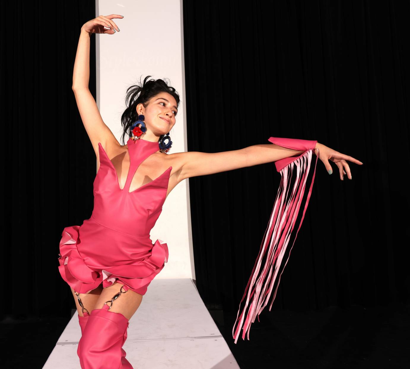 A woman in a red dress lifts her arms in an L. She looks to the left. A red bracelet with long fringe is on left wrist.